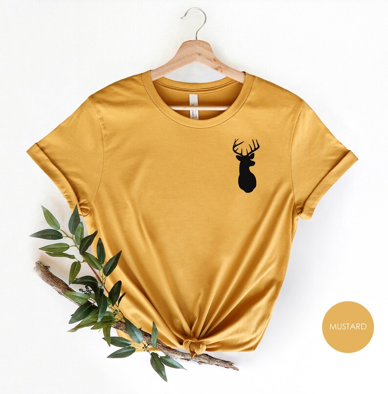 May include: A mustard yellow t-shirt with a black silhouette of a deer head on the left side of the chest. The shirt is on a hanger and has a knotted bottom.