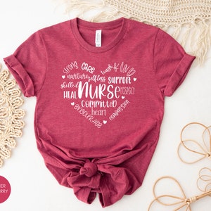 May include: Heather raspberry coloured t-shirt with white text that reads "Strong Care Tough Kind Nurturing Selfless Support Skilled Nurse Respect Heal Committed Heart Encourage Compassion".