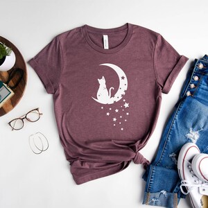 May include: A mauve t-shirt with a white graphic of a cat sitting on a crescent moon with stars. The t-shirt is laid out on a white surface with a pair of jeans, a pair of white sneakers, and a wooden tray with a plant, a camera, and glasses.