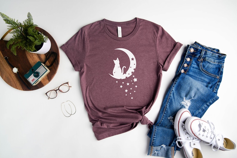 May include: A mauve t-shirt with a white graphic of a cat sitting on a crescent moon with stars. The t-shirt is laid out on a white surface with a pair of jeans, a pair of white sneakers, and a wooden tray with a plant, a camera, and glasses.