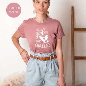 Puede incluir: Una mujer lleva una camiseta de color malva claro con el texto "Just a girl who loves chickens" y una silueta blanca de un pollo. Lleva pantalones cortos de mezclilla azul claro y un cintur&oacute;n marr&oacute;n.