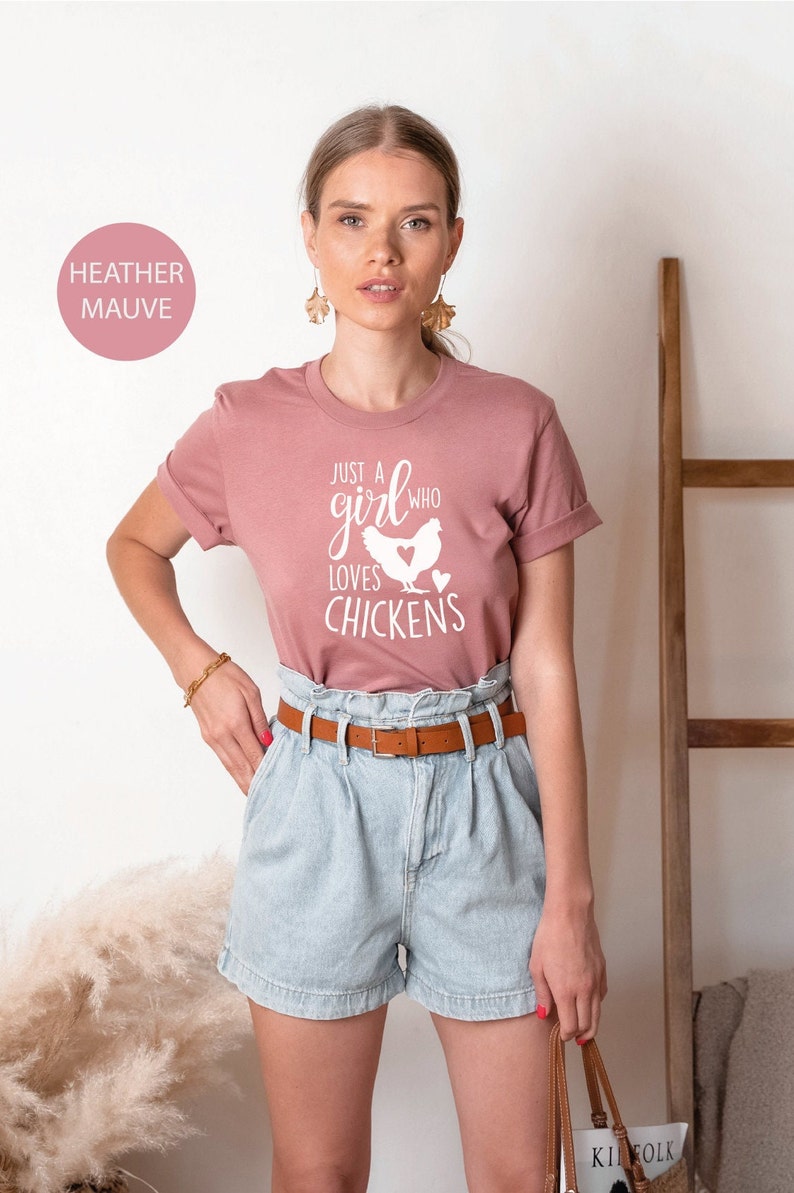 Puede incluir: Una mujer lleva una camiseta de color malva claro con el texto "Just a girl who loves chickens" y una silueta blanca de un pollo. Lleva pantalones cortos de mezclilla azul claro y un cintur&oacute;n marr&oacute;n.