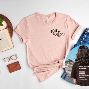 May include: A pink heather peach t-shirt with the text "You matter" printed on the front. The shirt is shown on a white background with a book, glasses, and a brown leather wallet. A second image shows a black t-shirt with the text "To the person behind me: You are amazing, beautiful and enough. Remember that." printed on the back.