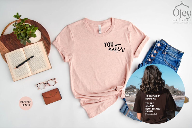May include: A pink heather peach t-shirt with the text "You matter" printed on the front. The shirt is shown on a white background with a book, glasses, and a brown leather wallet. A second image shows a black t-shirt with the text "To the person behind me: You are amazing, beautiful and enough. Remember that." printed on the back.