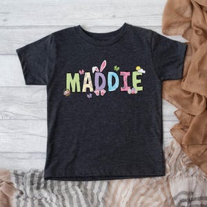 May include: Dark gray t-shirt with the name "MADDIE" in colorful, Easter-themed lettering. The design includes bunny ears, butterflies, and Easter eggs. The shirt is short-sleeved and appears to be made of a soft material.
