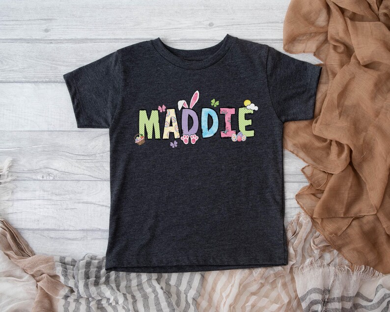 May include: Dark gray t-shirt with the name "MADDIE" in colorful, Easter-themed lettering. The design includes bunny ears, butterflies, and Easter eggs. The shirt is short-sleeved and appears to be made of a soft material.