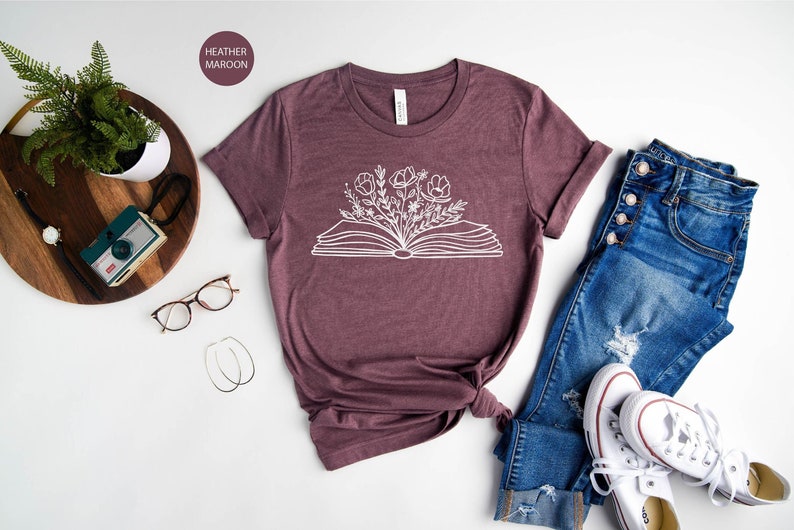 K&ouml;nnte beinhalten: Ein T-Shirt in Heide-Maroon mit einer wei&szlig;en Grafik eines offenen Buches, aus dem Blumen wachsen. Das Shirt liegt auf einer wei&szlig;en Oberfl&auml;che mit einer Brille, einer Kamera, einer Pflanze, einer Jeans und wei&szlig;en Turnschuhen.