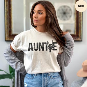 May include: Ivory-colored t-shirt with the word "AUNTIE" in bold, black letters. The letter "E" is replaced with a black skeleton hand making a rock-and-roll hand gesture. The model is wearing blue jeans and a gray cardigan.
