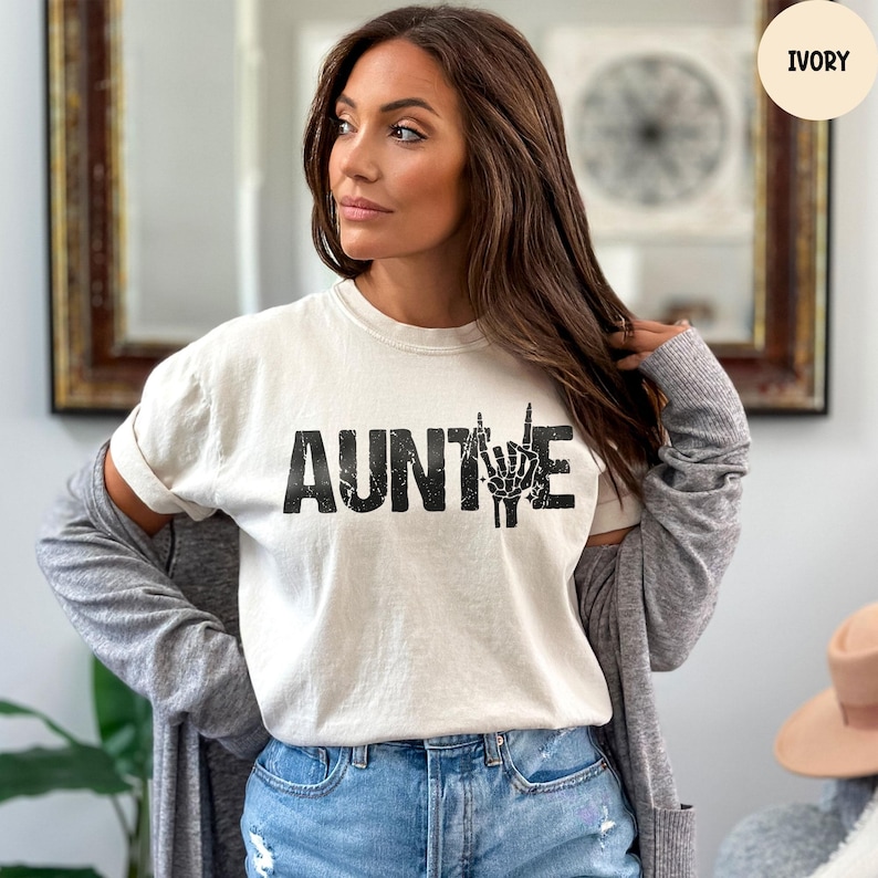 May include: Ivory-colored t-shirt with the word "AUNTIE" in bold, black letters. The letter "E" is replaced with a black skeleton hand making a rock-and-roll hand gesture. The model is wearing blue jeans and a gray cardigan.