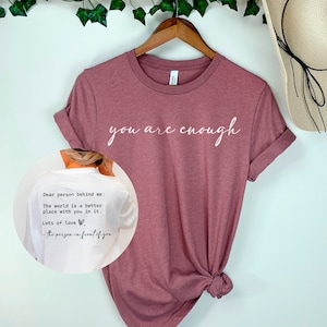 May include: A mauve t-shirt with the text "you are enough" printed in white on the front. The back of the shirt has a white note that reads "Dear person behind me: The world is a better place with you in it. Lots of love, - the person in front of you."