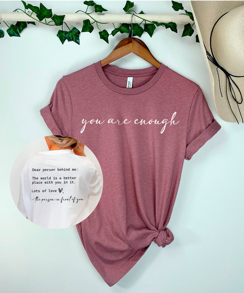 May include: A mauve t-shirt with the text "you are enough" printed in white on the front. The back of the shirt has a white note that reads "Dear person behind me: The world is a better place with you in it. Lots of love, - the person in front of you."