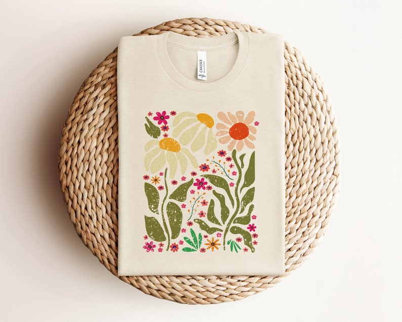 May include: A beige t-shirt with a floral print featuring white, pink, orange, and green flowers. The flowers are stylized and have a vintage feel.