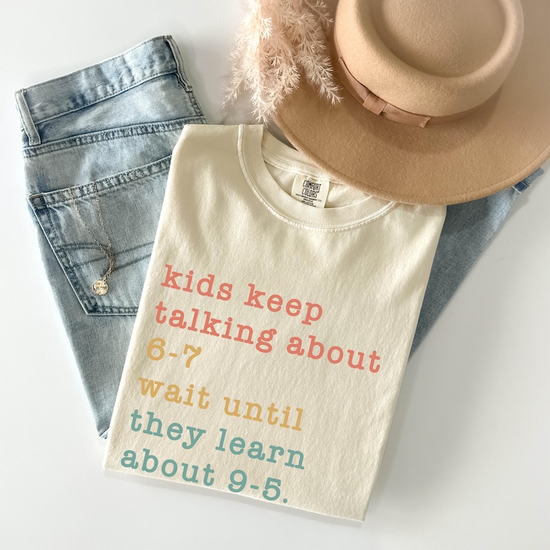 May include: Cream-coloured t-shirt with the text "kids keep talking about 6-7 wait until they learn about 9-5" in pink, yellow, and blue. The shirt is paired with light-wash jeans, a tan hat, and a necklace.
