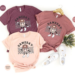 May include: Three t-shirts with the text "GAMEDAY" in a retro font. The shirts are in heather maroon, heather mauve, and heather peach. Each shirt features a cartoon baseball with a smiling face wearing a baseball cap.