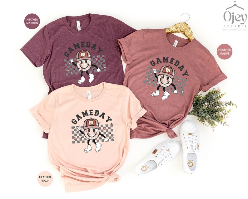 May include: Three t-shirts with the text "GAMEDAY" in a retro font. The shirts are in heather maroon, heather mauve, and heather peach. Each shirt features a cartoon baseball with a smiling face wearing a baseball cap.