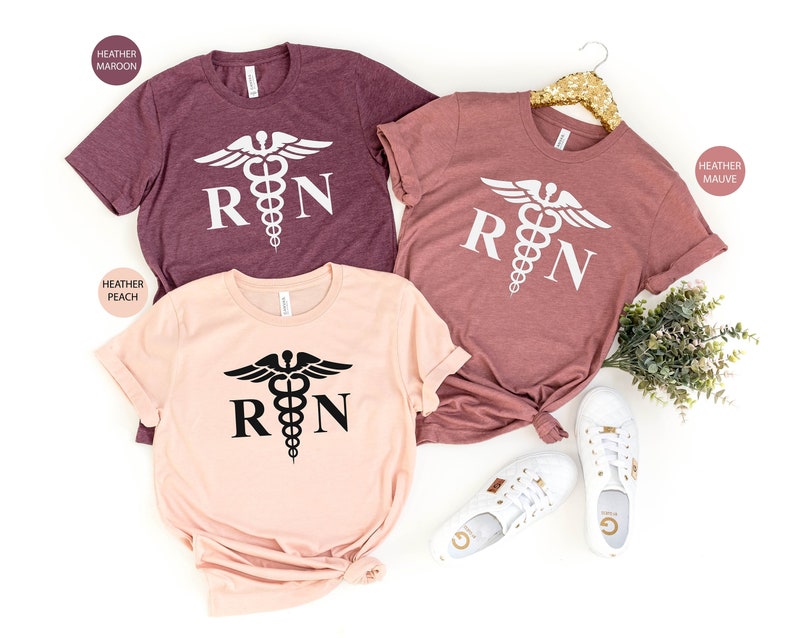 May include: Three t-shirts with the text "RN" and a medical symbol. The shirts are in heather maroon, heather peach, and heather mauve.