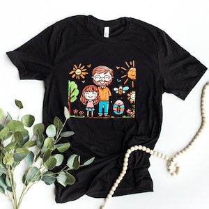 Peut inclure: T-shirt noir avec une illustration color&eacute;e d'un p&egrave;re et de sa fille dans un jardin. L'illustration est dessin&eacute;e dans un style enfantin avec des couleurs vives et des lignes simples.