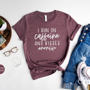 K&ouml;nnte beinhalten: Heather-marronfarbenes T-Shirt mit dem Text "I run on caffeine and kisses #momlife" in Wei&szlig;. Das Shirt ist auf einer wei&szlig;en Oberfl&auml;che mit einer Brille, einem Holztablett mit einer Kamera und einer Pflanze, einer Jeans und wei&szlig;en Turnschuhen abgebildet.