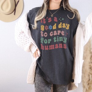 May include: A dark gray t-shirt with the text "It's a good day to care for tiny humans" in colorful retro lettering. The shirt is being worn by a person with a white cardigan.