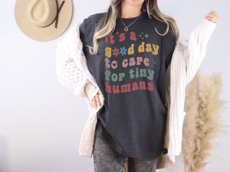 May include: A dark gray t-shirt with the text "It's a good day to care for tiny humans" in colorful retro lettering. The shirt is being worn by a person with a white cardigan.