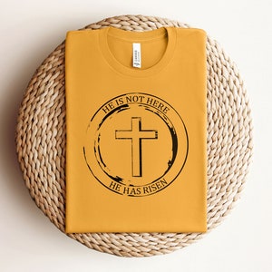 K&ouml;nnte beinhalten: Ein senfgelbes T-Shirt mit einem schwarzen Kreis-Motiv, das ein Kreuz und den Text "Er ist nicht hier, Er ist auferstanden" zeigt.