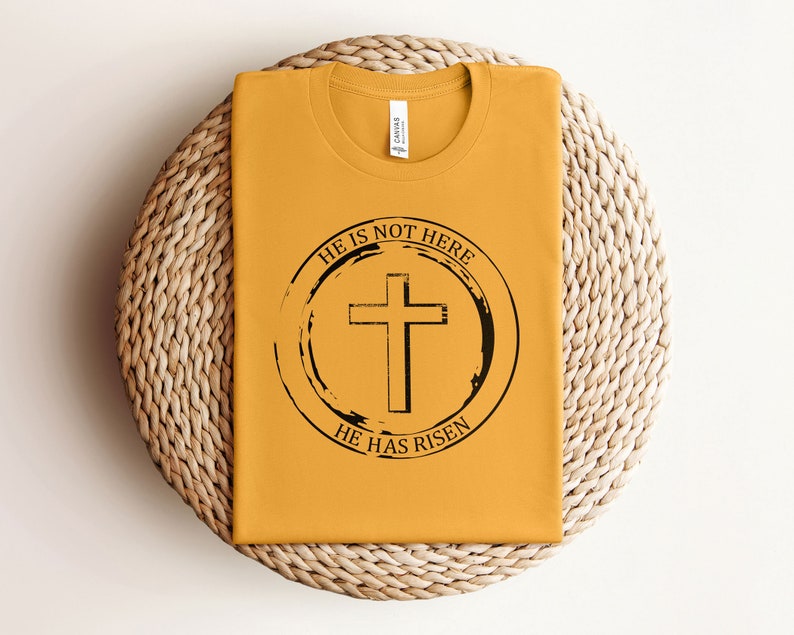 K&ouml;nnte beinhalten: Ein senfgelbes T-Shirt mit einem schwarzen Kreis-Motiv, das ein Kreuz und den Text "Er ist nicht hier, Er ist auferstanden" zeigt.