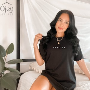 Op de afbeelding: Een zwarte t-shirtjurk met het woord "REALTOR" in wit op de voorkant. Het model draagt een gouden ketting.