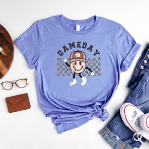 May include: A heather columbia blue t-shirt with a graphic of a smiling baseball wearing a hat and the text "GAMEDAY" in black letters.