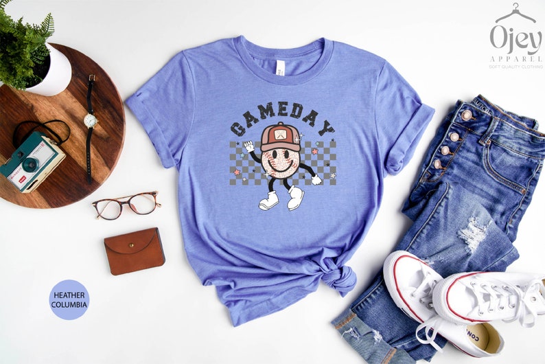 May include: A heather columbia blue t-shirt with a graphic of a smiling baseball wearing a hat and the text "GAMEDAY" in black letters.