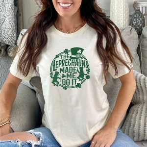 May include: A white t-shirt with a green graphic design that says "The Leprechauns Made Me Do It". The graphic features two leprechauns, shamrocks, and a pot of gold.