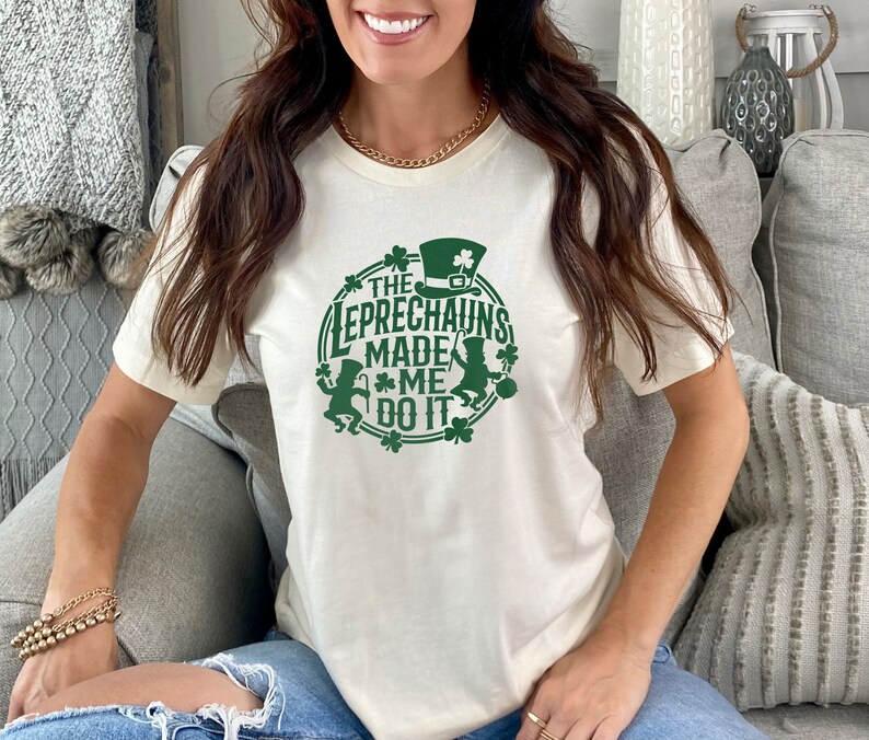 May include: A white t-shirt with a green graphic design that says "The Leprechauns Made Me Do It". The graphic features two leprechauns, shamrocks, and a pot of gold.