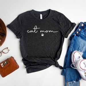 May include: Heather dark gray t-shirt with the text "cat mom" in white script with a paw print. The shirt is laid flat on a white surface with a pair of jeans, a purse, and a potted plant.