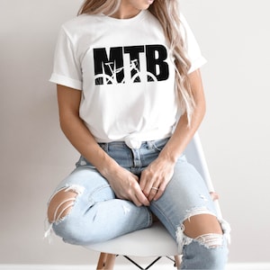 Puede incluir: Una camiseta blanca con un gr&aacute;fico negro de una bicicleta de monta&ntilde;a y el texto "MTB". La persona que lleva la camiseta est&aacute; sentada en una silla blanca y lleva pantalones vaqueros azules.