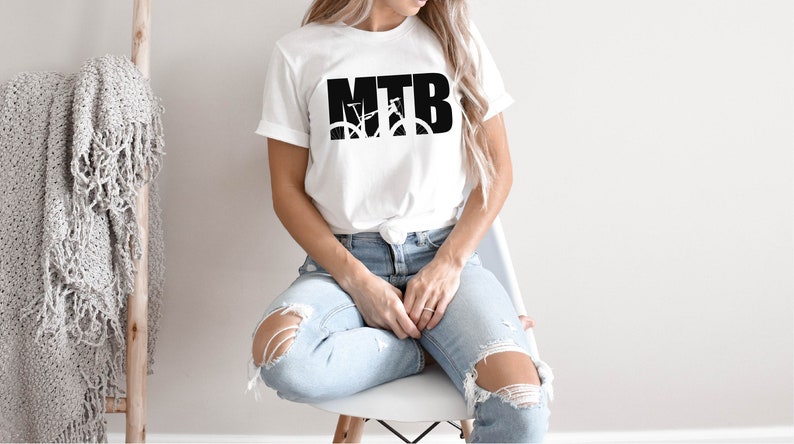 Puede incluir: Una camiseta blanca con un gr&aacute;fico negro de una bicicleta de monta&ntilde;a y el texto "MTB". La persona que lleva la camiseta est&aacute; sentada en una silla blanca y lleva pantalones vaqueros azules.