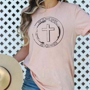 K&ouml;nnte beinhalten: Ein rosa T-Shirt mit einem schwarzen Kreis-Motiv, das ein Kreuz und den Text "Er ist nicht hier, Er ist auferstanden" zeigt.
