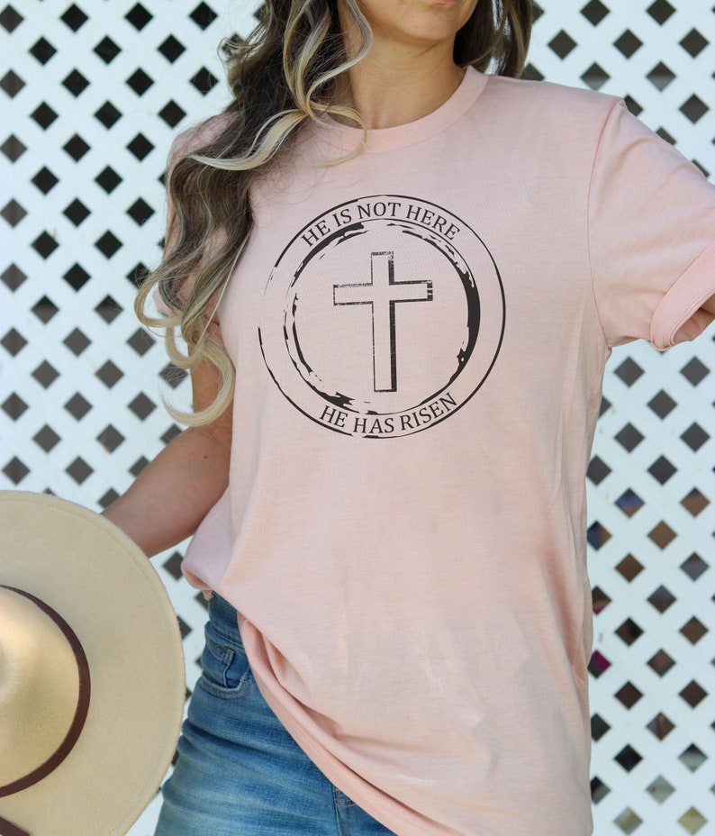 K&ouml;nnte beinhalten: Ein rosa T-Shirt mit einem schwarzen Kreis-Motiv, das ein Kreuz und den Text "Er ist nicht hier, Er ist auferstanden" zeigt.