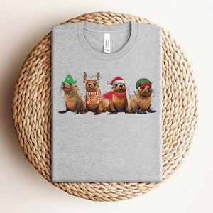 May include: A gray t-shirt with a Christmas-themed design featuring four sea lions wearing festive hats and decorations. The sea lions are wearing Santa hats, reindeer antlers, and a green Christmas hat. They are surrounded by Christmas lights.