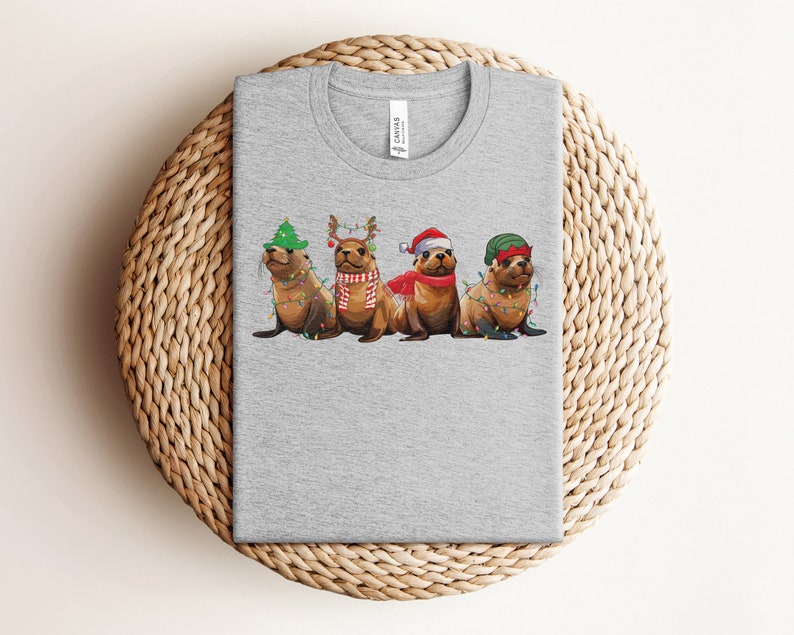 May include: A gray t-shirt with a Christmas-themed design featuring four sea lions wearing festive hats and decorations. The sea lions are wearing Santa hats, reindeer antlers, and a green Christmas hat. They are surrounded by Christmas lights.