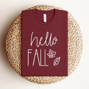 May include: A maroon t-shirt with the text "hello fall" in white lettering and a white outline of a maple leaf and an oak leaf.