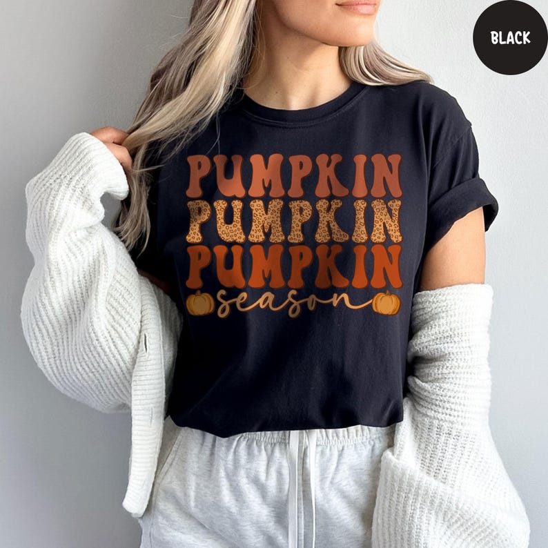 May include: Black t-shirt with the words "PUMPKIN PUMPKIN PUMPKIN season" in orange and leopard print lettering. Two orange pumpkins are on either side of the word "season". The shirt is a fall-themed graphic tee.