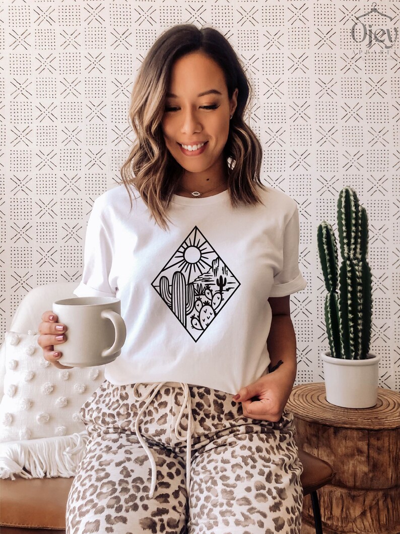May include: White t-shirt with a black graphic of a desert landscape with a sun, mountains, and cacti. The graphic is inside a diamond shape.