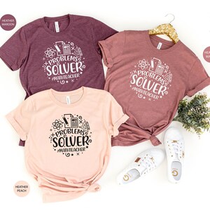 May include: Three t-shirts with the text "Problems Solver #MathTeacher" in a circle with math symbols. The shirts are in heather maroon, heather peach, and heather mauve.