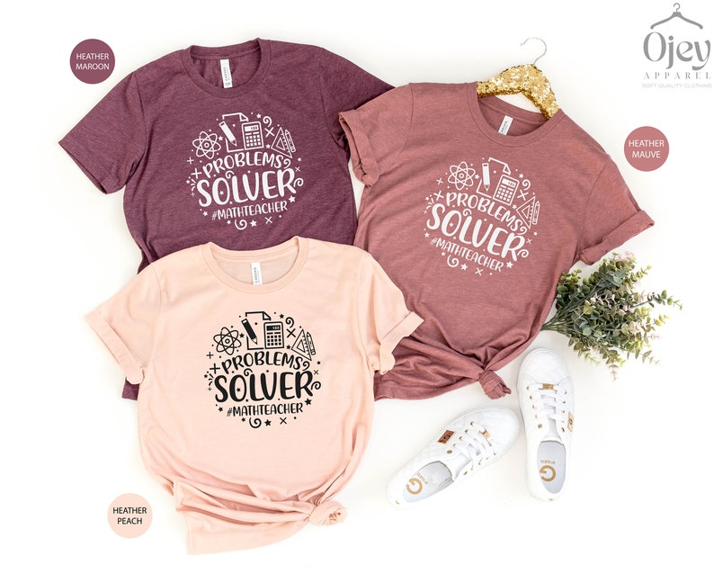 May include: Three t-shirts with the text "Problems Solver #MathTeacher" in a circle with math symbols. The shirts are in heather maroon, heather peach, and heather mauve.