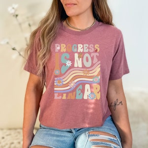 May include: A mauve cropped t-shirt with a graphic print that reads "Progress is not linear" in a retro font with colorful flowers and wavy lines.