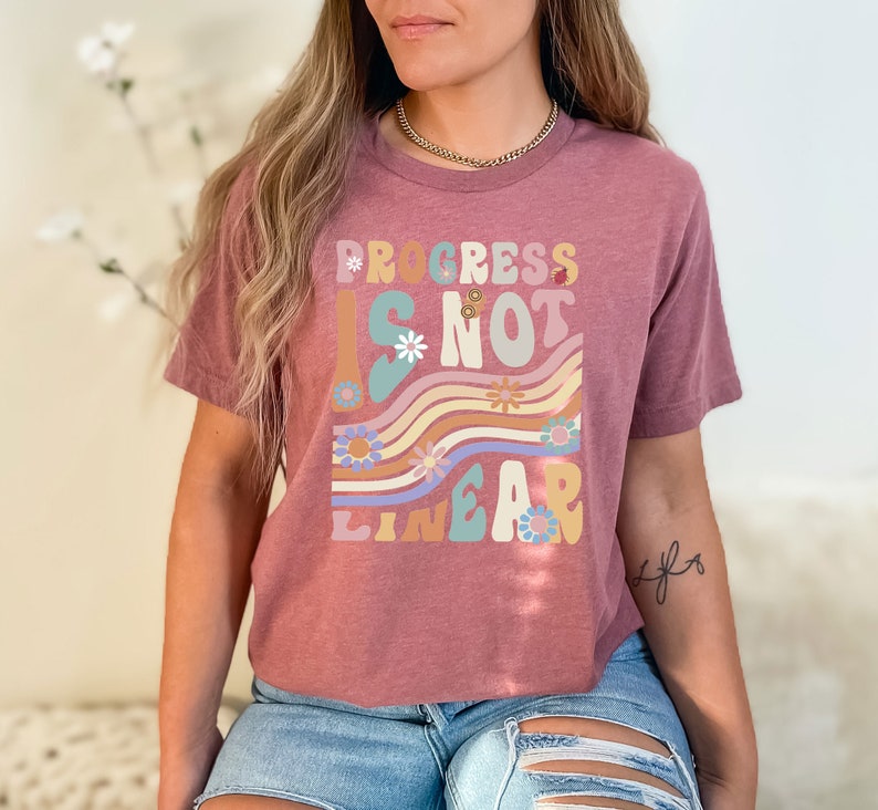 May include: A mauve cropped t-shirt with a graphic print that reads "Progress is not linear" in a retro font with colorful flowers and wavy lines.