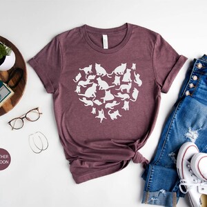 May include: A heather maroon t-shirt with a white graphic of a heart made up of cats. The shirt is laid out on a white surface with a pair of glasses, a wooden tray with a camera, and a pair of jeans and white sneakers.