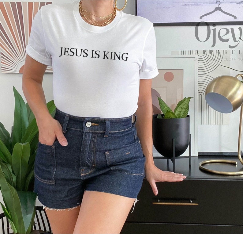 Pode incluir: Camiseta branca com o texto "JESUS IS KING" impresso em preto. A camiseta est&aacute; sendo usada por uma pessoa que est&aacute; usando shorts jeans azuis.