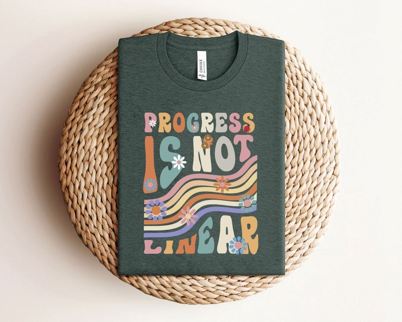 May include: A dark green t-shirt with the text "Progress is not linear" in a retro, colorful font with flowers and wavy lines.