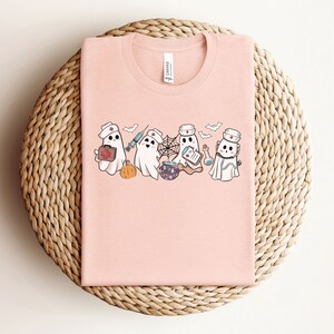 May include: Pink t-shirt featuring a design of four ghosts dressed as nurses. The ghosts are holding medical supplies such as a syringe, a first aid kit, a potion bottle, and a clipboard. The text "Boo Crew" is printed on the shirt.