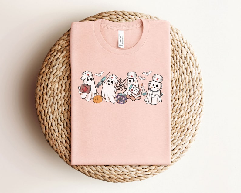 May include: Pink t-shirt featuring a design of four ghosts dressed as nurses. The ghosts are holding medical supplies such as a syringe, a first aid kit, a potion bottle, and a clipboard. The text "Boo Crew" is printed on the shirt.